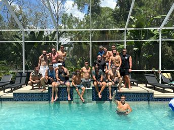 Group of men at a sunny backyard screened pool party, sitting on a tiled ledge and lounge chairs with lush tropical plants behind; one man in the water raises a drink.