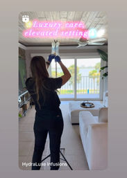 Clinician adjusting an IV infusion bag in a bright waterfront living room with white sectional sofa, wood‑paneled ceiling, ceiling fan and balcony palm‑tree view