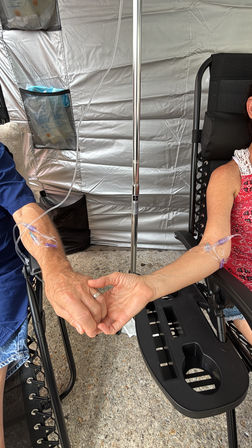 Two adults holding hands while each receives an IV drip in their forearms, seated in reclining chairs next to an IV pole inside a medical tent.
