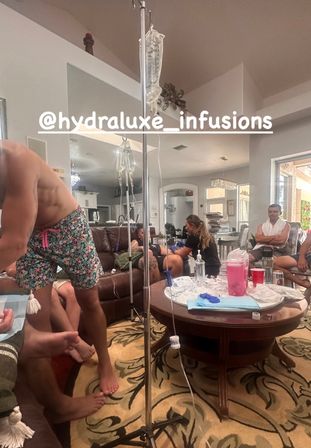 Cozy living-room at-home IV therapy session: IV pole with drip bags, clinician administering an infusion at a couch, coffee table strewn with medical supplies and people relaxing.