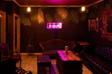 Speakeasy-style lounge with neon sign, dark palm-print wallpaper, tufted leather sofas and armchairs, wooden trunk coffee table with stacked wooden blocks, warm hanging bulbs and purple-pink mood lighting.