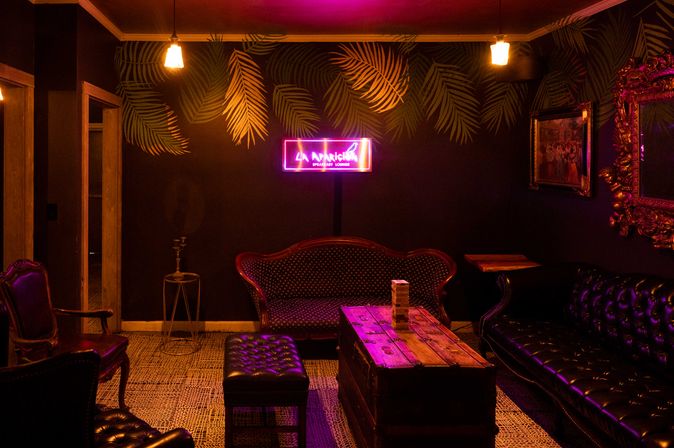 Speakeasy-style lounge with neon sign, dark palm-print wallpaper, tufted leather sofas and armchairs, wooden trunk coffee table with stacked wooden blocks, warm hanging bulbs and purple-pink mood lighting.