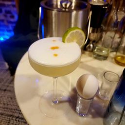 Frothy citrus sour cocktail in a coupe with lime wheel and three bitters drops on egg-white foam, served on a bar table next to an egg in a shot glass.
