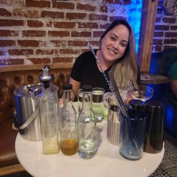 Smiling person seated at a round table in a brick-walled cocktail bar with glass bottles, shakers, and mixology tools on the table.