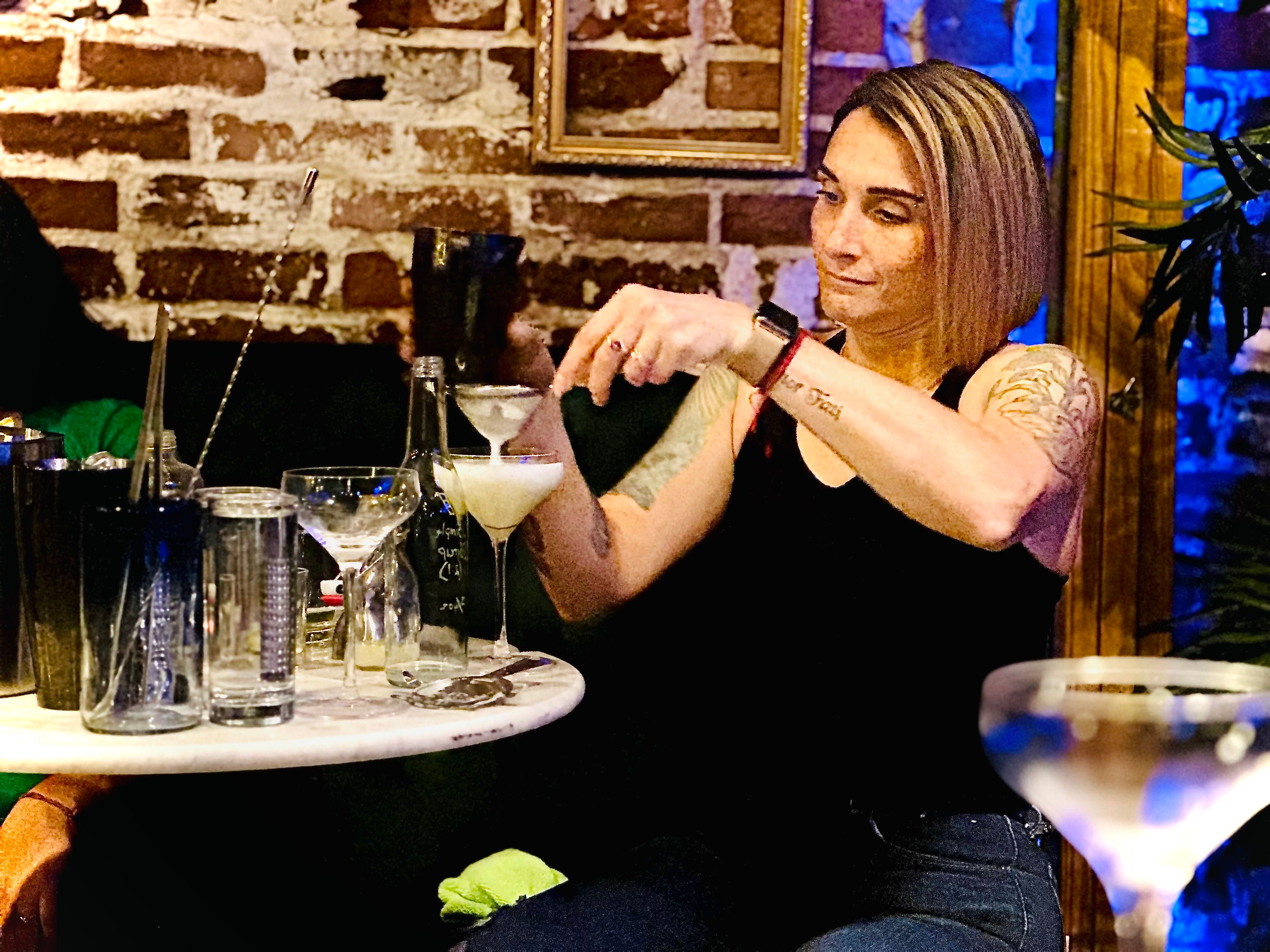 Bartender pouring a frothy cocktail into a coupe glass at a cozy brick-wall cocktail lounge, bar tools and glassware on a round marble table.