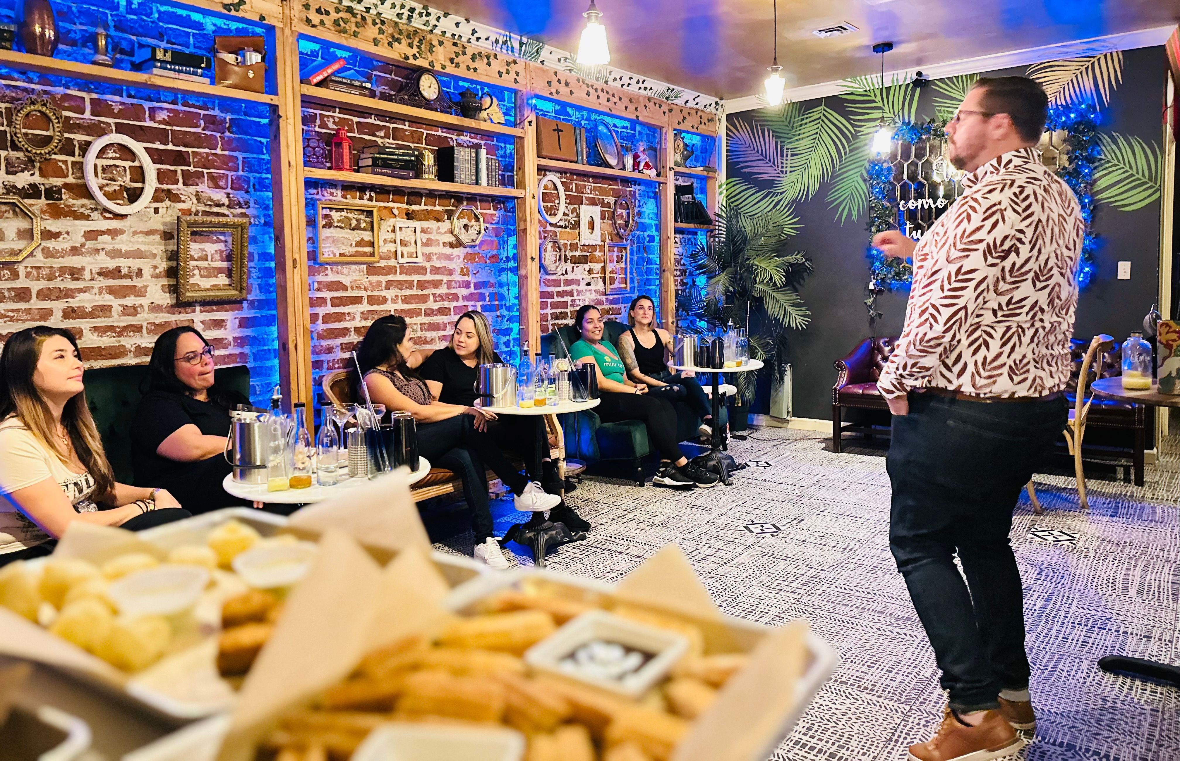 Presenter speaking to a seated group in a cozy lounge with exposed brick, blue accent lighting, small tables and snacks in the foreground during a cocktail-tasting style event.