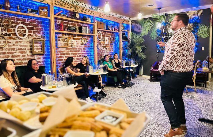 Presenter speaking to a seated group in a cozy lounge with exposed brick, blue accent lighting, small tables and snacks in the foreground during a cocktail-tasting style event.