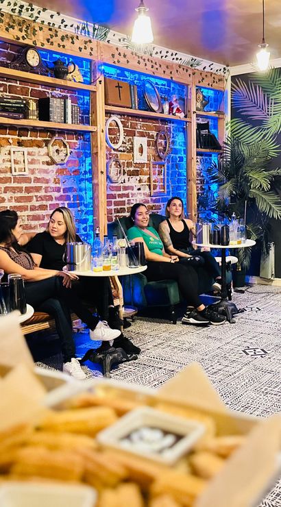 Presenter speaking to a seated group in a cozy lounge with exposed brick, blue accent lighting, small tables and snacks in the foreground during a cocktail-tasting style event.