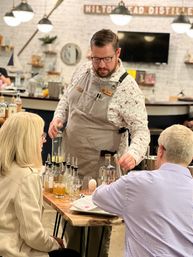 Mixologist in an apron pouring spirits into glasses at a wooden table during a craft distillery tasting, with small spirit bottles, cocktail tools and two seated guests in a cozy tasting room with pendant lights.