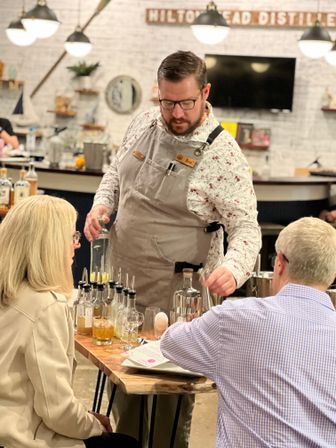 Mixologist in an apron pouring spirits into glasses at a wooden table during a craft distillery tasting, with small spirit bottles, cocktail tools and two seated guests in a cozy tasting room with pendant lights.
