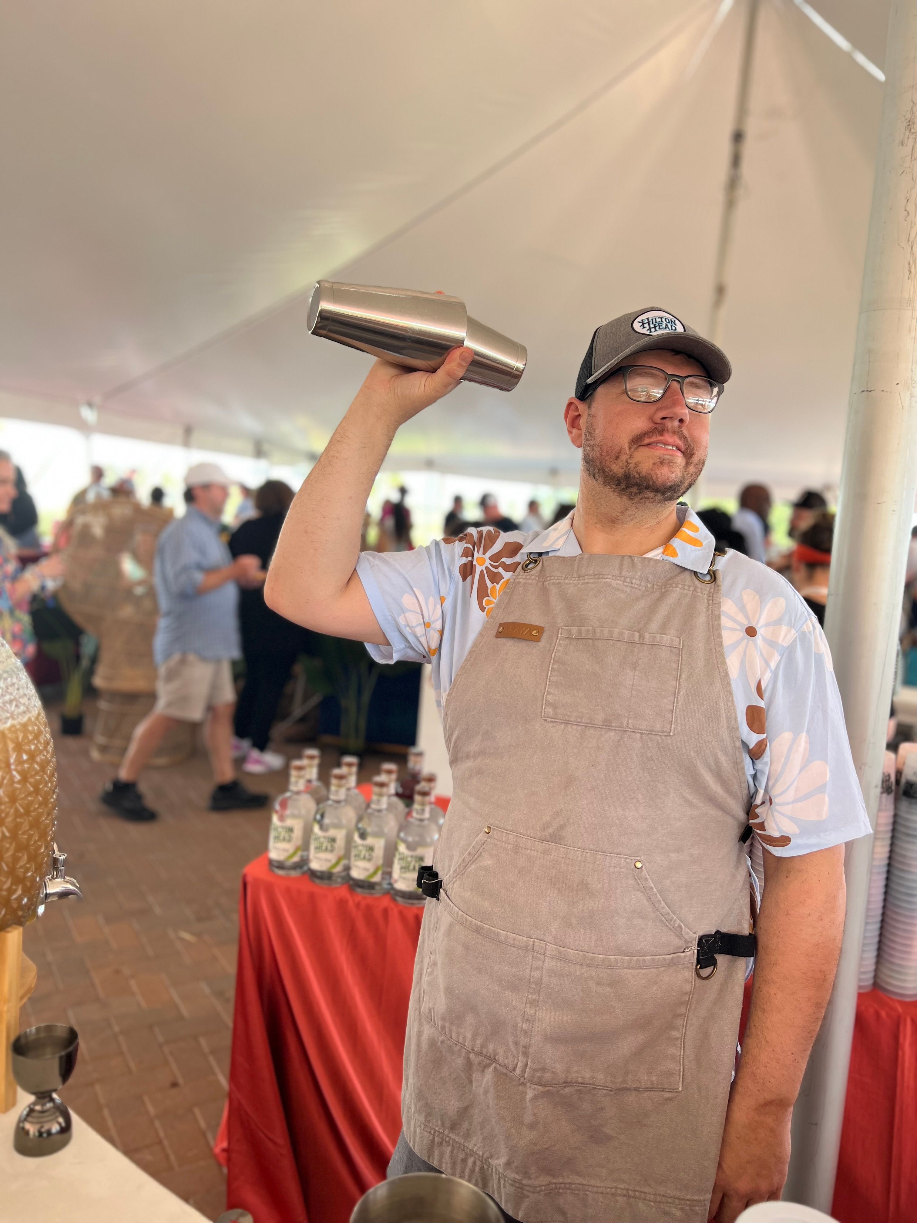 Bartender in a floral shirt and apron shaking a stainless cocktail shaker inside a crowded outdoor tasting tent at a food festival, with bottles lined on a red-covered table.
