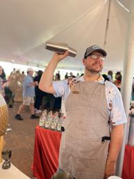 Bartender in a floral shirt and apron shaking a stainless cocktail shaker inside a crowded outdoor tasting tent at a food festival, with bottles lined on a red-covered table.