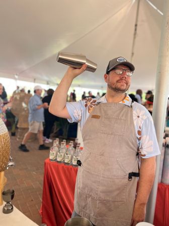 Bartender in a floral shirt and apron shaking a stainless cocktail shaker inside a crowded outdoor tasting tent at a food festival, with bottles lined on a red-covered table.