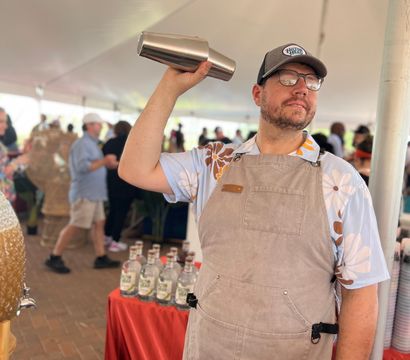Bartender in a floral shirt and apron shaking a stainless cocktail shaker inside a crowded outdoor tasting tent at a food festival, with bottles lined on a red-covered table.