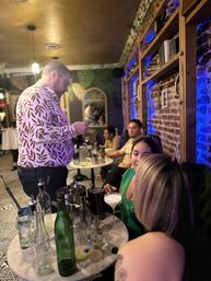 Friends enjoying cocktails at an intimate downtown lounge — bartender in a patterned shirt pouring drinks at a small marble table with bottles, brick wall shelves and blue accent lighting.
