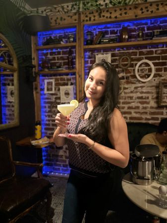 Smiling woman holding a lime‑garnished margarita in a cozy brick‑walled bar with blue LED shelves and vintage decor.