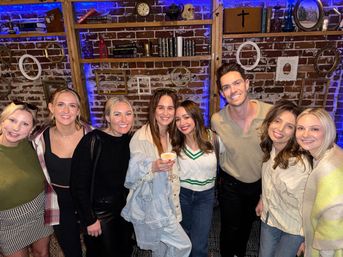 Cheerful group of eight friends smiling for a night-out photo in a cozy brick-walled lounge with blue accent lighting, vintage shelves and one person holding a cocktail.