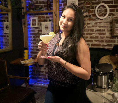 Person holding a lime-garnished cocktail in a cozy brick-walled cocktail bar with blue accent lighting and vintage wall frames.