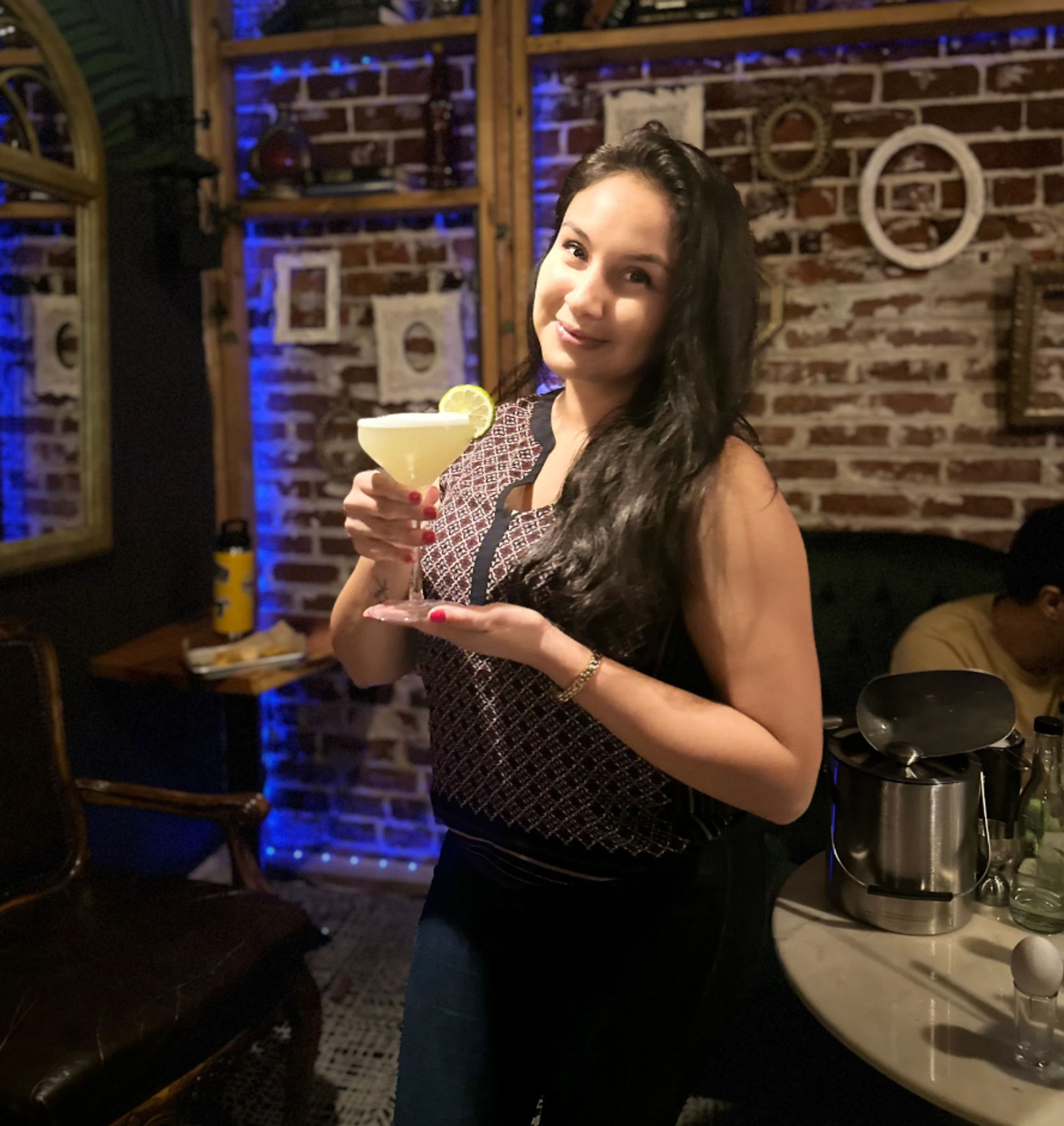 Person holding a lime-garnished cocktail in a cozy brick-walled cocktail bar with blue accent lighting and vintage wall frames.