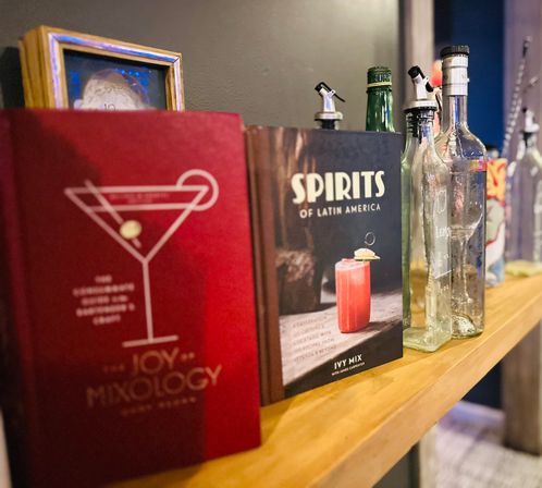 Wooden bar shelf with cocktail books (one titled "Spirits of Latin America"), assorted glass liquor bottles with pour spouts and a playful mixology vibe.