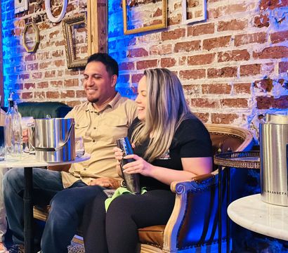 Two people laughing on a vintage leather sofa in a cozy brick-walled cocktail bar, one holding a cocktail shaker with metal ice buckets, glass bottles and a marble table nearby, decorative empty frames and blue accent lighting on the wall.