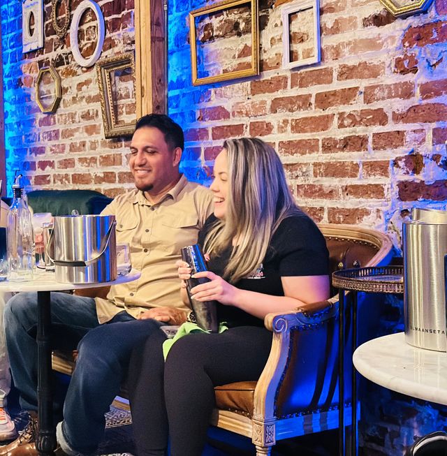 Two people laughing on a vintage leather sofa in a cozy brick-walled cocktail bar, one holding a cocktail shaker with metal ice buckets, glass bottles and a marble table nearby, decorative empty frames and blue accent lighting on the wall.