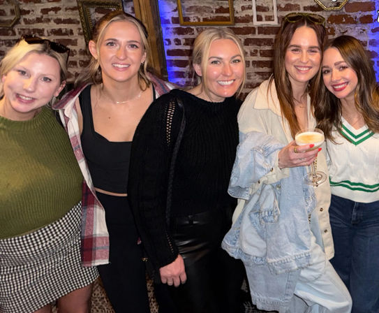 Five friends smiling and posing in a cozy brick-walled bar, one holding a cocktail glass — casual jackets and sweaters, lively night-out vibe.