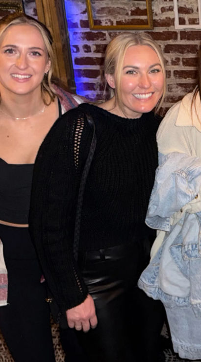 Five friends smiling and posing in a cozy brick-walled bar, one holding a cocktail glass — casual jackets and sweaters, lively night-out vibe.