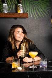 Smiling female bartender holding a tall citrus cocktail at a stylish tropical-themed cocktail bar, colorful mixed drinks on a black marble counter