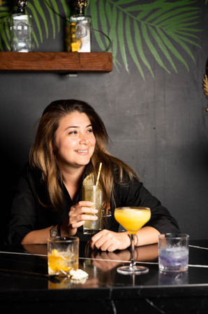 Smiling female bartender holding a tall citrus cocktail at a stylish tropical-themed cocktail bar, colorful mixed drinks on a black marble counter