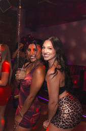 Two smiling women posing on a nightclub dance floor under red disco lights — one holding a cocktail in a purple dress, the other in a black top with a leopard-print skirt, partying with friends.