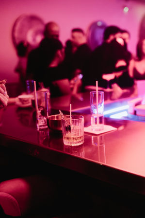 Neon-pink bar counter scattered with cocktail glasses, straws and a small bowl, blurred crowd mingling in the background — lively nightlife scene.