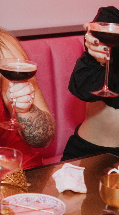 Group of friends toasting with coupe cocktails in a pink cocktail lounge booth, smiling women in a red dress and black crop top and assorted drinks on the table.