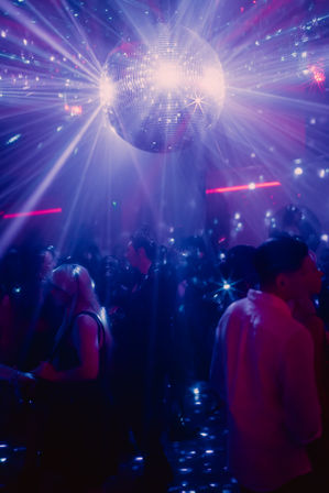 Mirrored disco ball sending purple and blue light rays across a crowded nightclub dance floor as partygoers dance under sparkling beams.