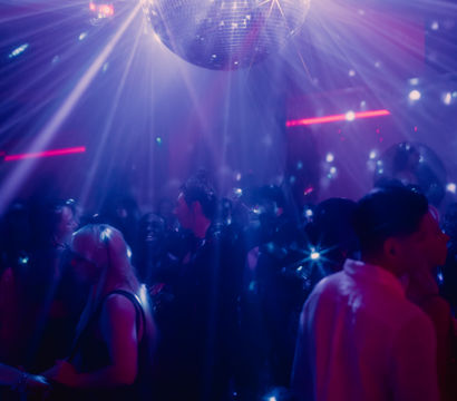 Mirrored disco ball sending purple and blue light rays across a crowded nightclub dance floor as partygoers dance under sparkling beams.