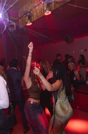 Two people dancing with cocktails under a disco ball on a red-lit nightclub dance floor, crowded nightlife scene with party lighting and upbeat atmosphere
