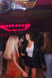 Two friends laughing and dancing on a crowded nightclub dance floor under a mirror disco ball and red circular LED lights