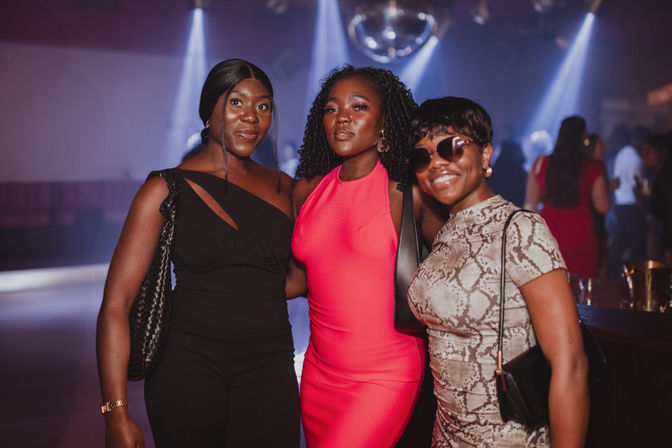 Three stylish women in cocktail attire striking a pose under a disco ball and colorful lights in a lively nightclub