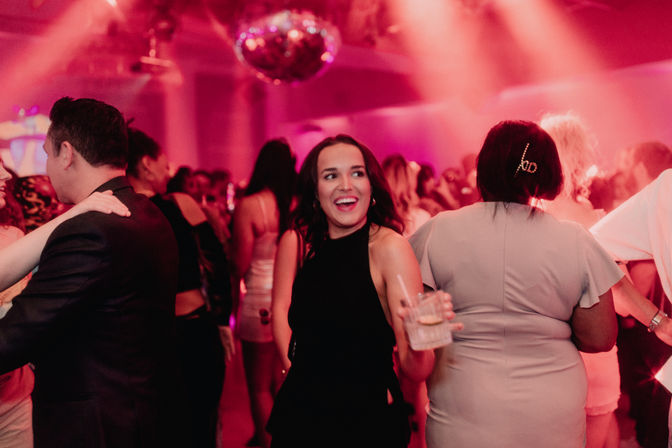 Woman in a black dress holding a drink and smiling on a crowded dance floor under a disco ball with pink club lighting, lively nightlife scene