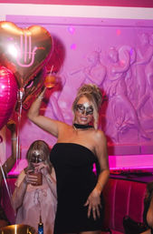 Masked woman in a black strapless dress holding a cocktail and heart-shaped balloons in a vibrant pink lounge with classical wall reliefs — masquerade girls' night out.