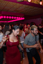 Vibrant nightclub dance floor with pink globe lights, crowded bar scene, woman in a fitted red dress holding a wine glass and a smiling man with a drink.