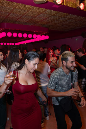 Vibrant nightclub dance floor with pink globe lights, crowded bar scene, woman in a fitted red dress holding a wine glass and a smiling man with a drink.
