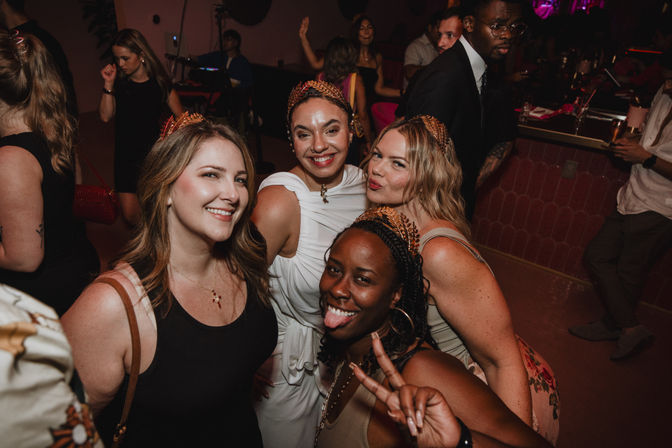 Four friends wearing decorative crowns smile and pose at a lively indoor nightlife party near a dimly lit bar, one flashing a peace sign and sticking out her tongue.