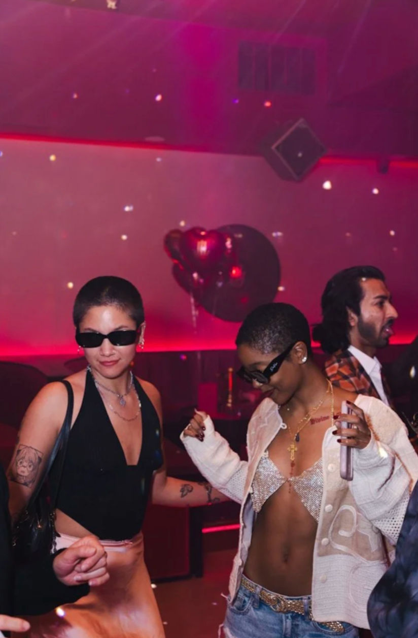 Two people dancing in a pink-lit nightclub — one in a black top and sunglasses, the other in a sparkly bralette, cardigan and jeans — with heart-shaped balloons and disco lights