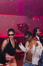 Two people dancing in a pink-lit nightclub — one in a black top and sunglasses, the other in a sparkly bralette, cardigan and jeans — with heart-shaped balloons and disco lights