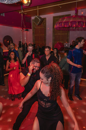 Energetic nightclub dance floor with a diverse crowd of young adults dancing under red lighting and a disco ball, DJ booth and ornate lounge decor