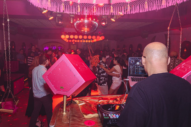 DJ at a crowded urban nightclub booth overlooking a packed dance floor with a disco ball, pink lighting, and people dancing