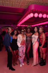 Group of six women posing in a pink-lit nightclub under rows of round globe lights, wearing party outfits—mini dresses, ripped jeans, crop tops and heels—smiling in an urban nightlife scene.