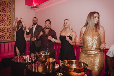 Five partygoers in a pink-lit VIP lounge, dressed for a night out — women in black and gold dresses and two men in smart-casual jackets — holding cocktails around tables with gold ice buckets and glassware.