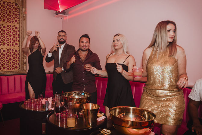 Five partygoers in a pink-lit VIP lounge, dressed for a night out — women in black and gold dresses and two men in smart-casual jackets — holding cocktails around tables with gold ice buckets and glassware.